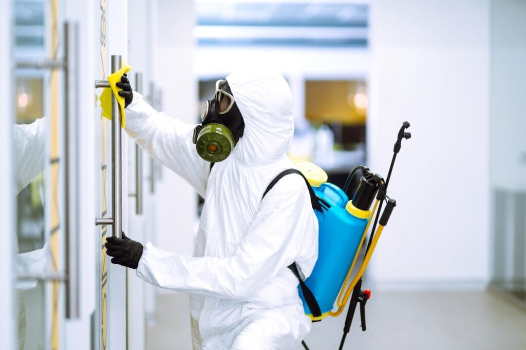 Man in protective hazmat suit disinfects office space to prevent COVID-19, emphasizing safety and cleanliness measures.